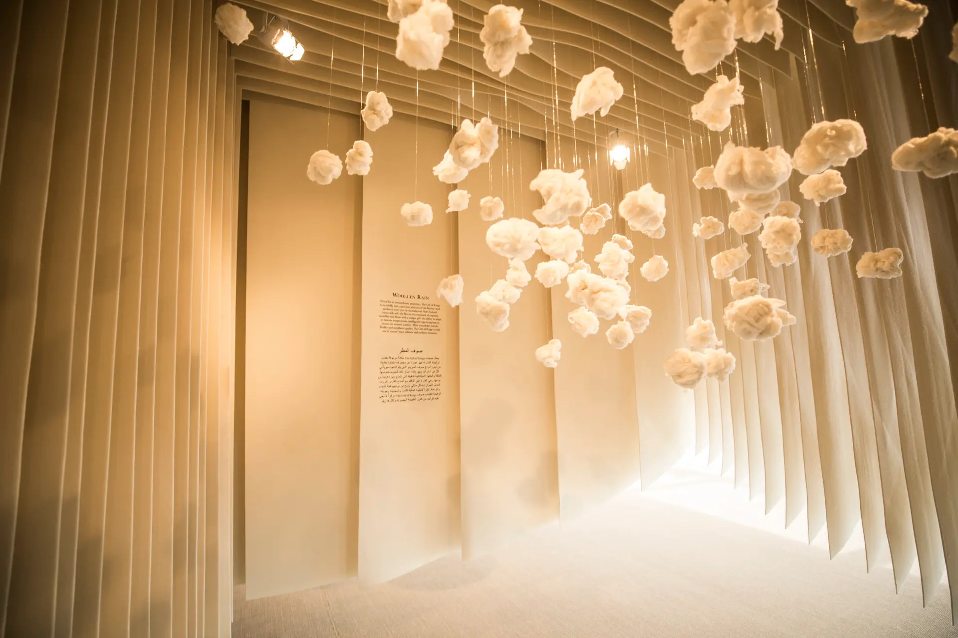 An immersive installation space with soft, hanging wool forms suspended from the ceiling, creating a cloud-like effect within a sculpted, light-filled room.